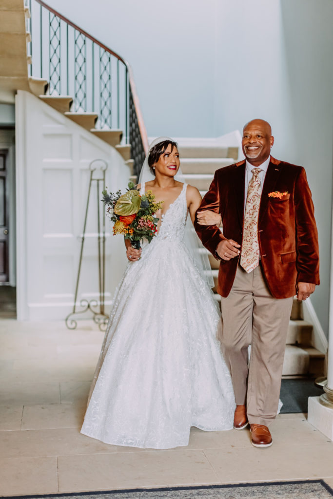 Bride and her dad walking through the Garthmyl Hall hallway with the staircase behind them