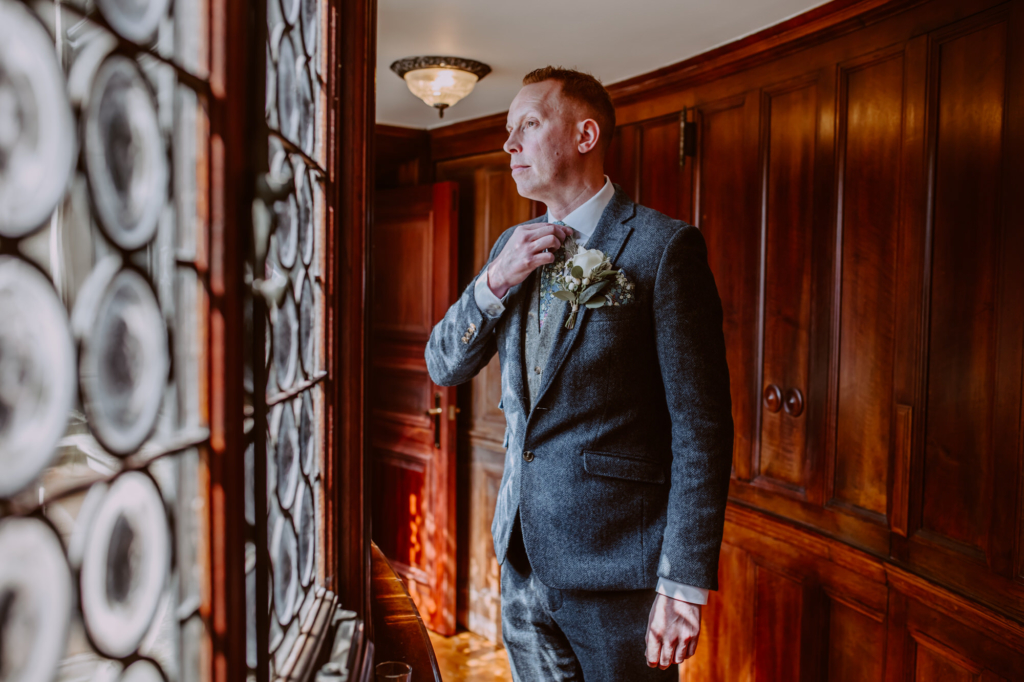 Groom looking out the window posing for portrait in corridor leading to the chapel
