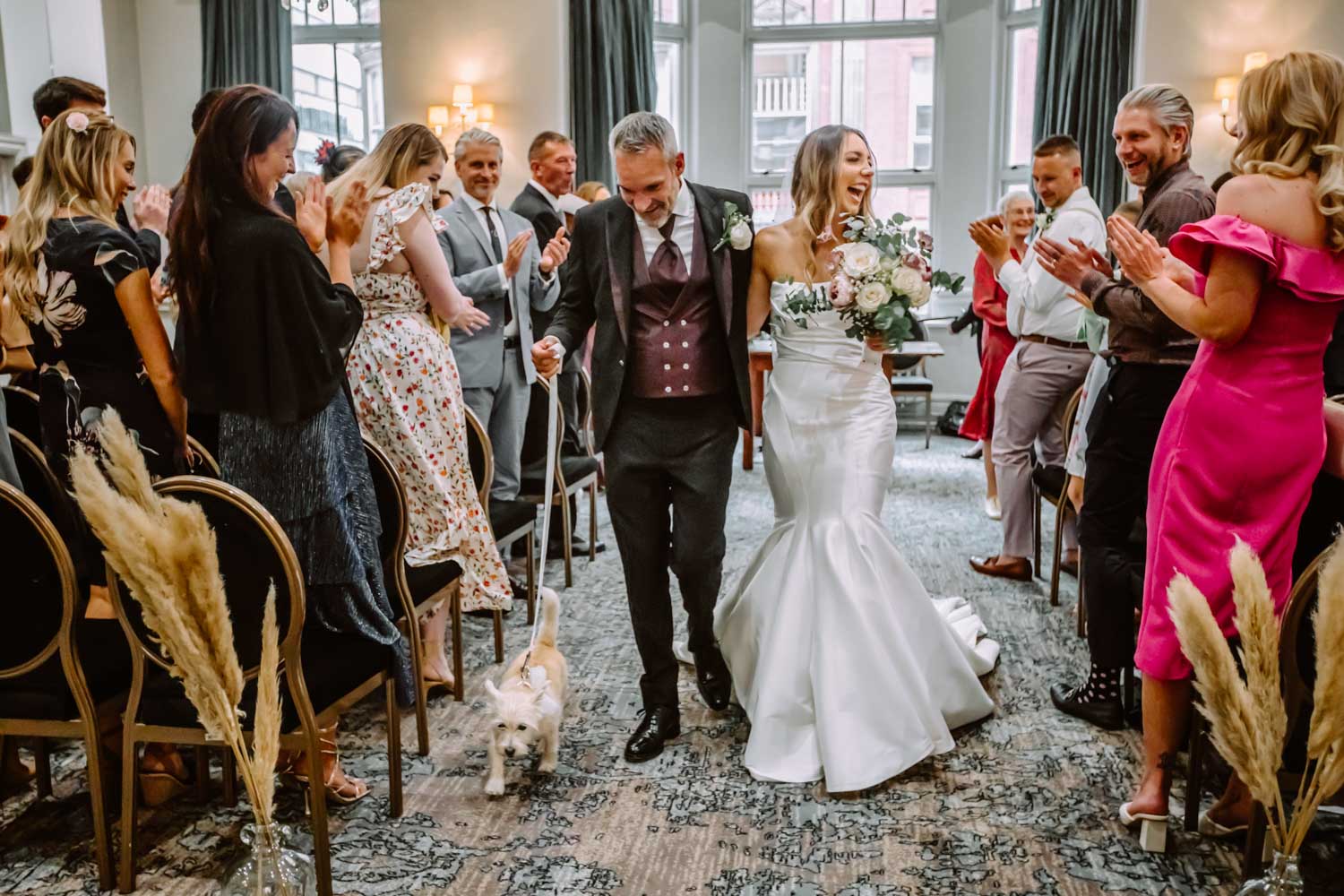 The freshly married couple joyfully walks down the aisle with their dog while guests clapping 