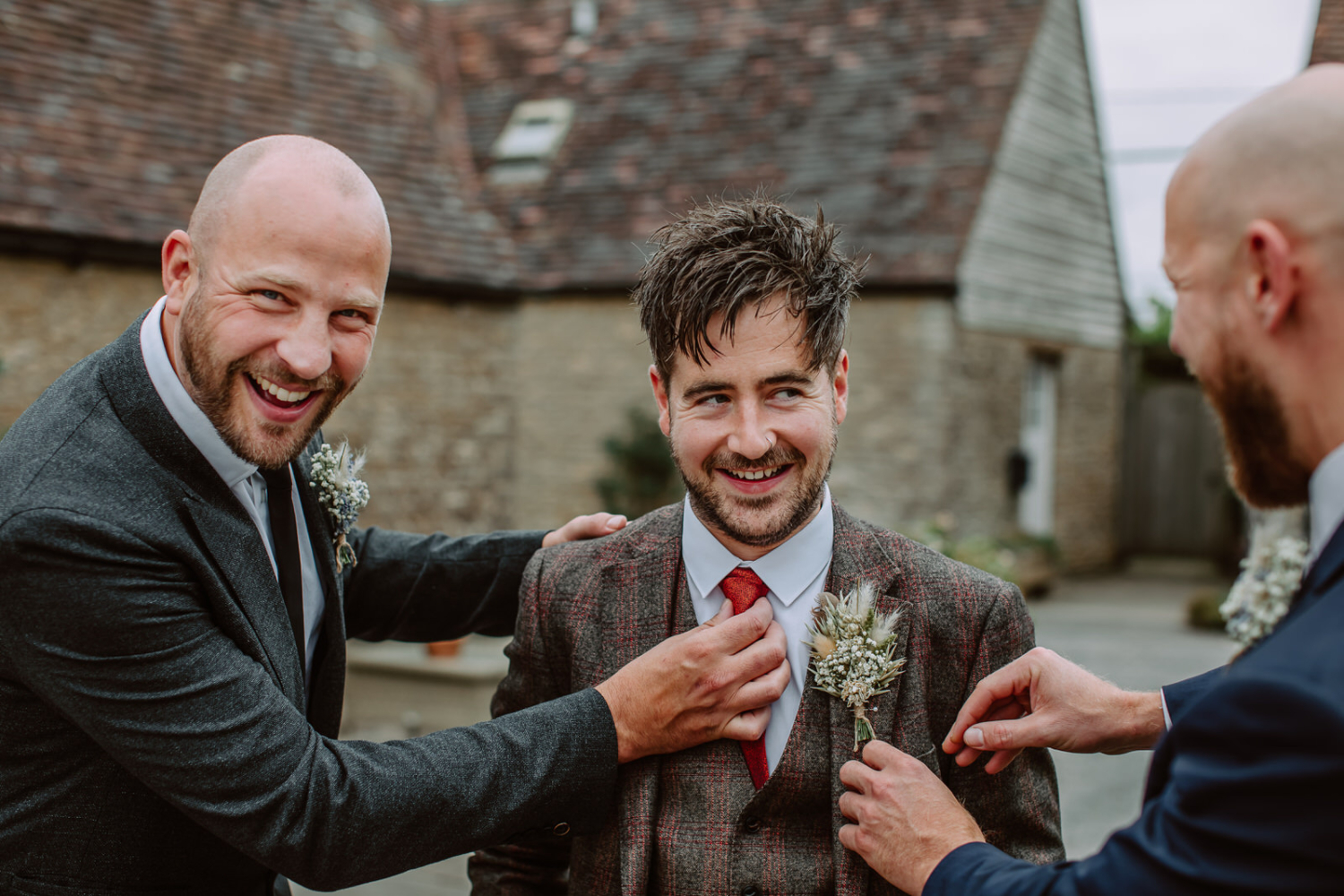 Fund photo of groomsman adjusting grooms tie and buttonhole while laughing.