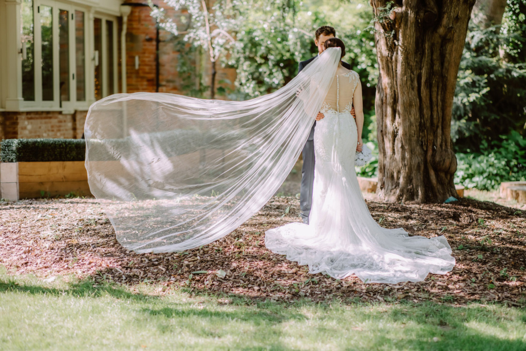 A bride and groom standing under a tree with their veil blowing in the wind.