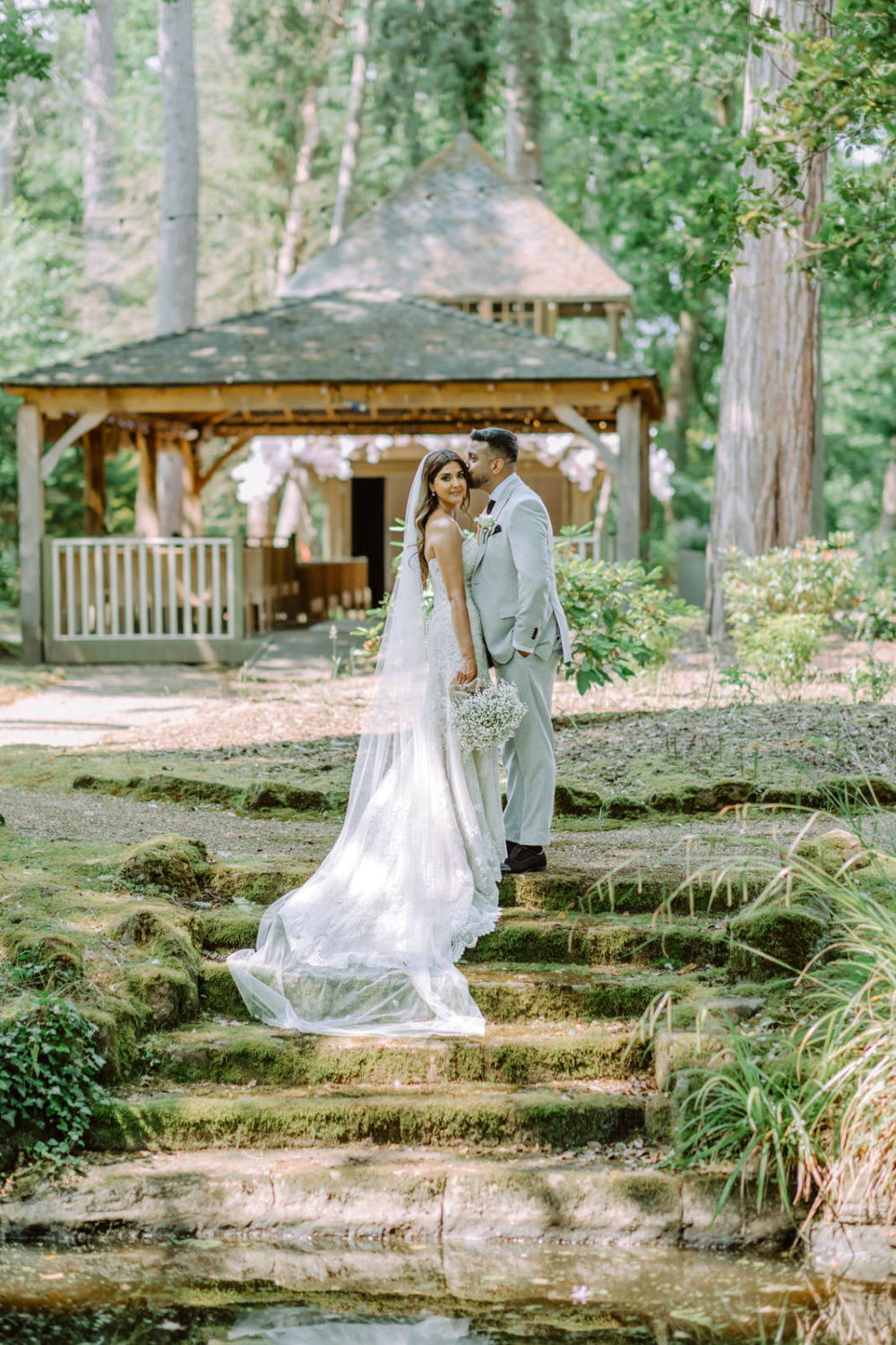 A bride and groom standing on steps near a pond at Hogarths Hotel Solihull on The Island surrounded by wooden grounds.