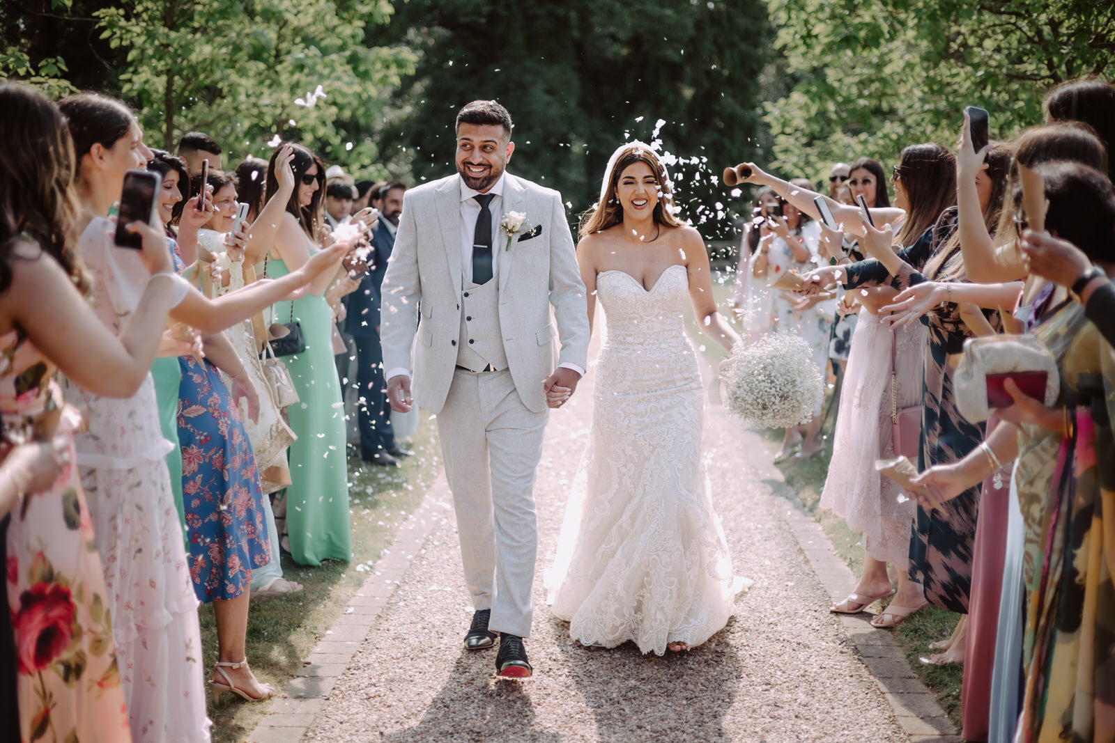 A bride and groom walk down an outdoor path, smiling, as guests on either side throw confetti and take photos with their phones.