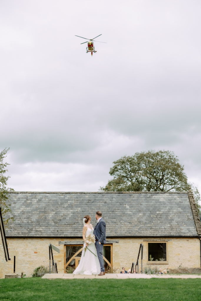 A bride and groom standing in front of a barn with a helicopter flying overhead.