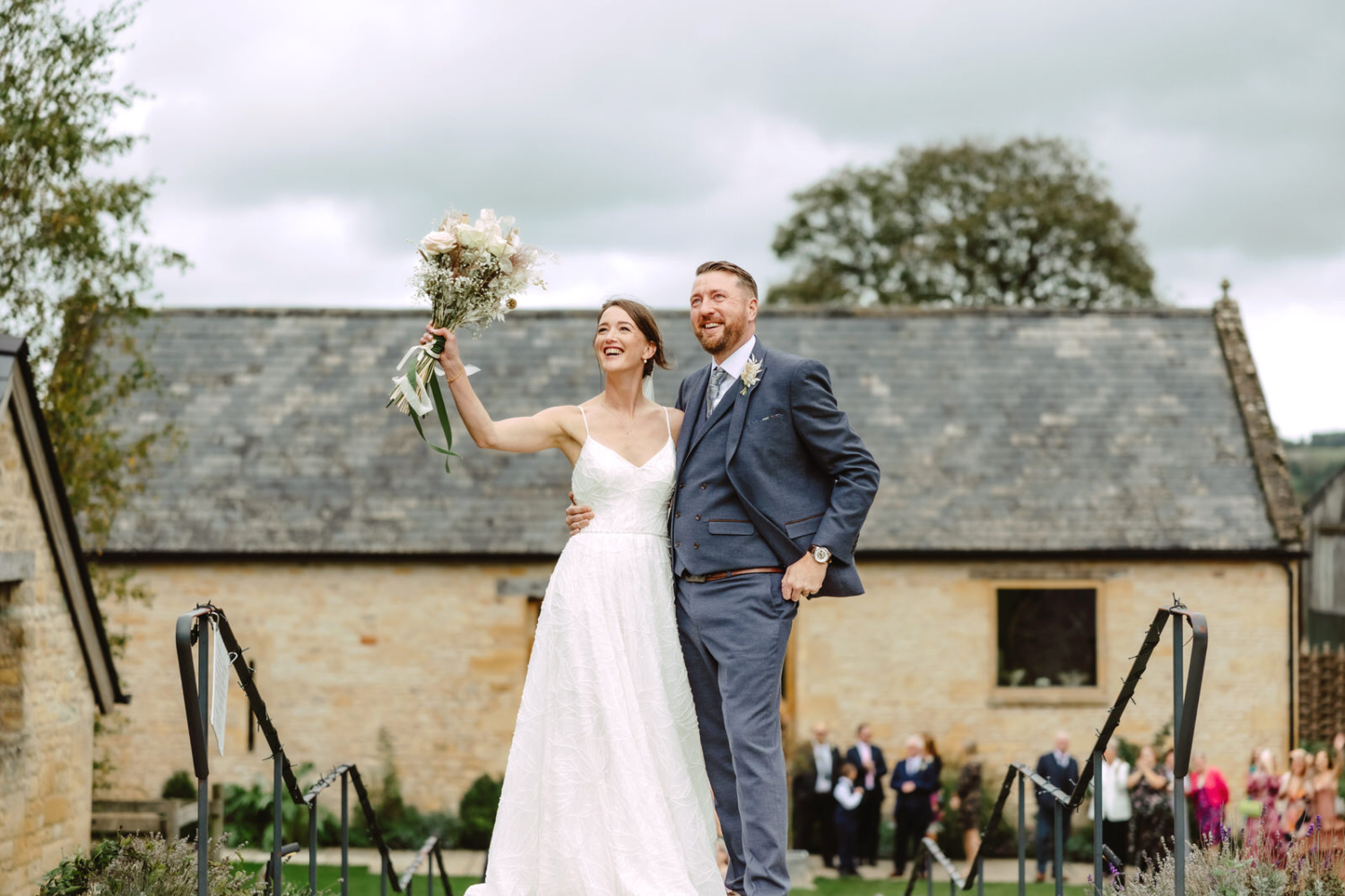 A bride and groom standing on the steps of their wedding venue. The bride is waving with her bouquet on a helicopter