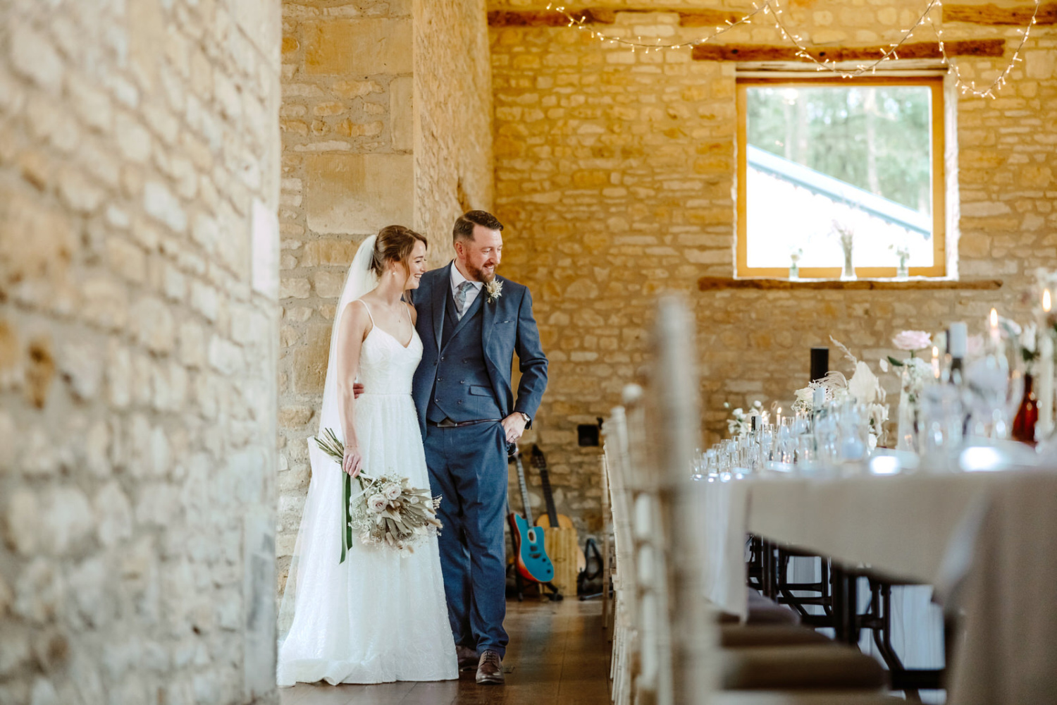 A bride and groom standing in a stone barn admiring the wedding breakfast set up.