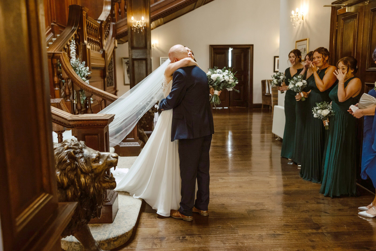 A bride and father of bride hugging in front of a staircase.
