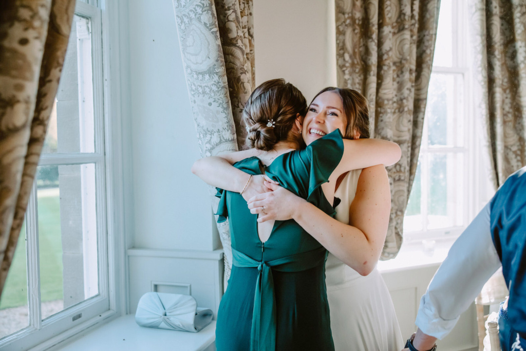 A bride hugging her bridesmaid in front of a window.