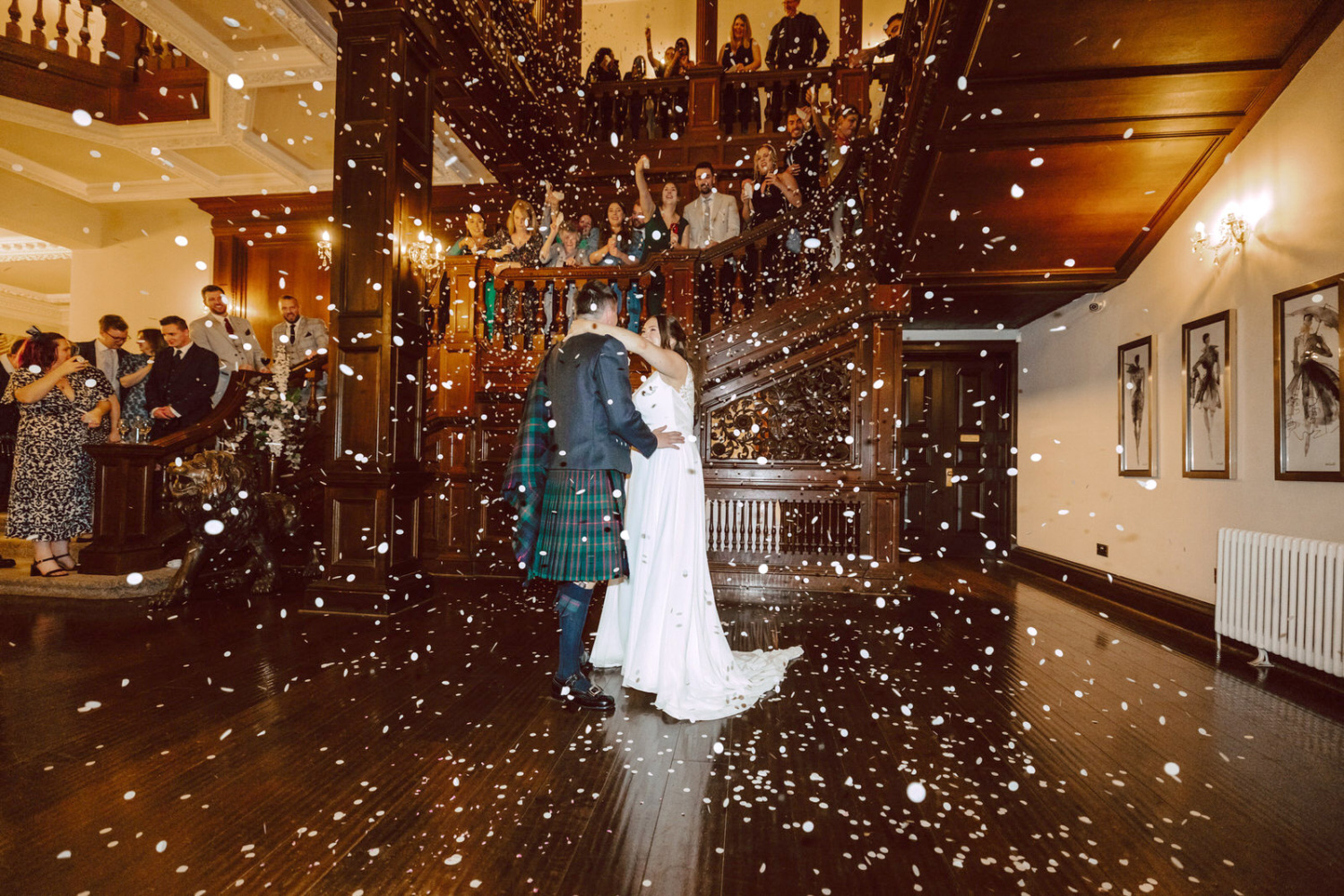 A bride and groom kissing in front of confetti. Guest are on the staircase of Bourton Hall
