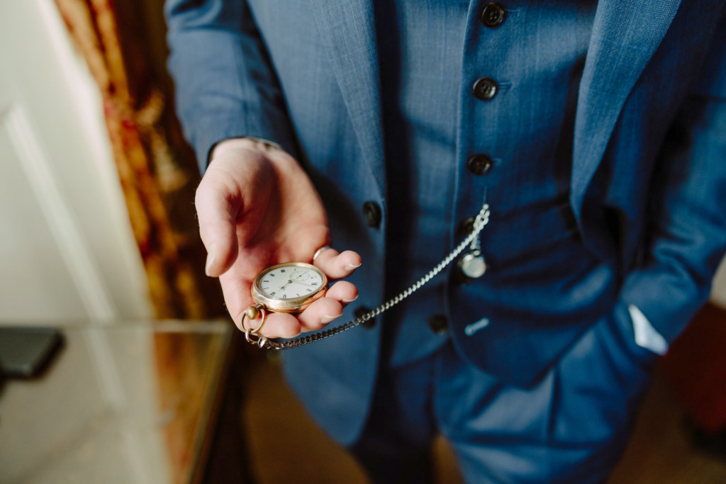 A groom in a blue suit holding a pocket watch.