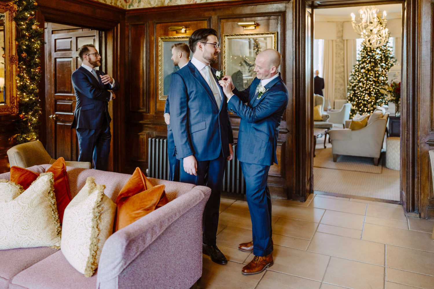 The groom place a buttonhole for his best man in a suit is getting ready in a room with a christmas tree at Rowton castle hallway.