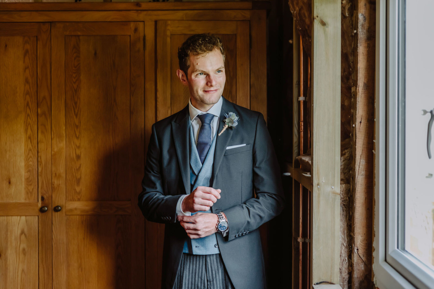 A groom in a suit standing in front of a window.