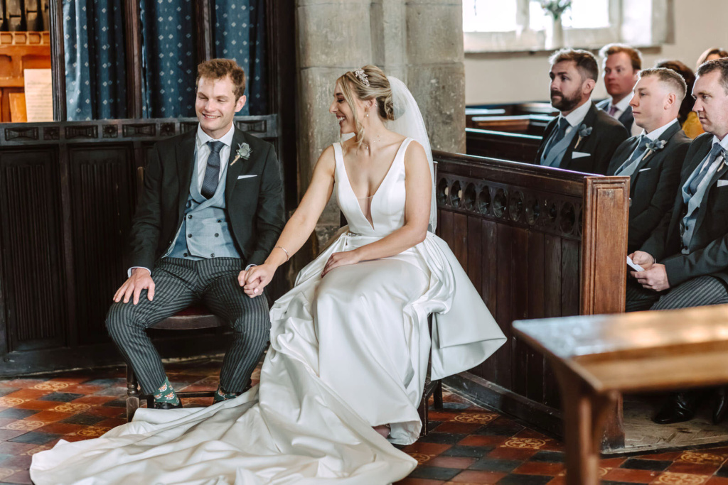 A bride and groom sitting in the pews of a church holding hands.