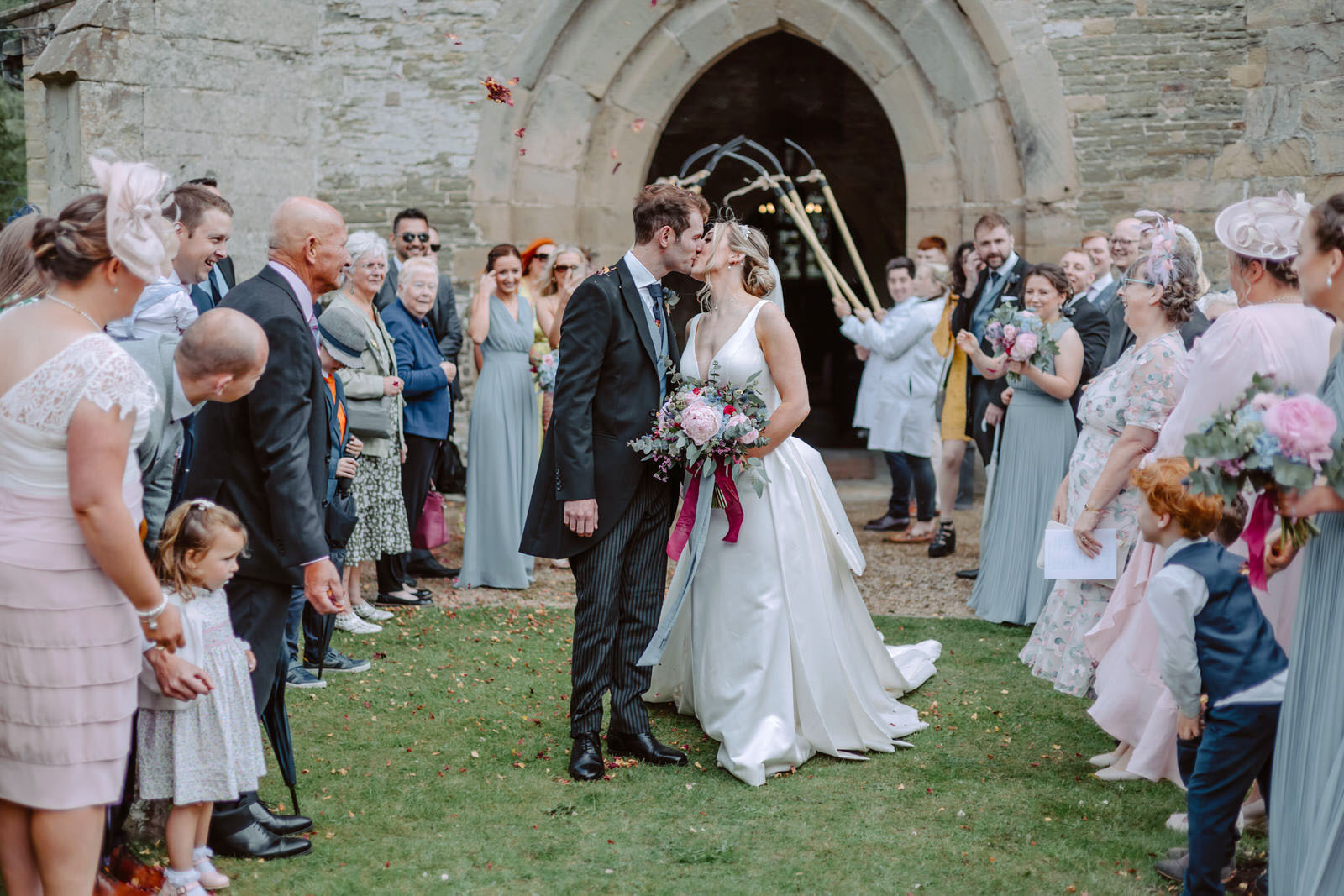 The bride and the groom kiss in front of the church surrounded by the wedding guests smiling. 