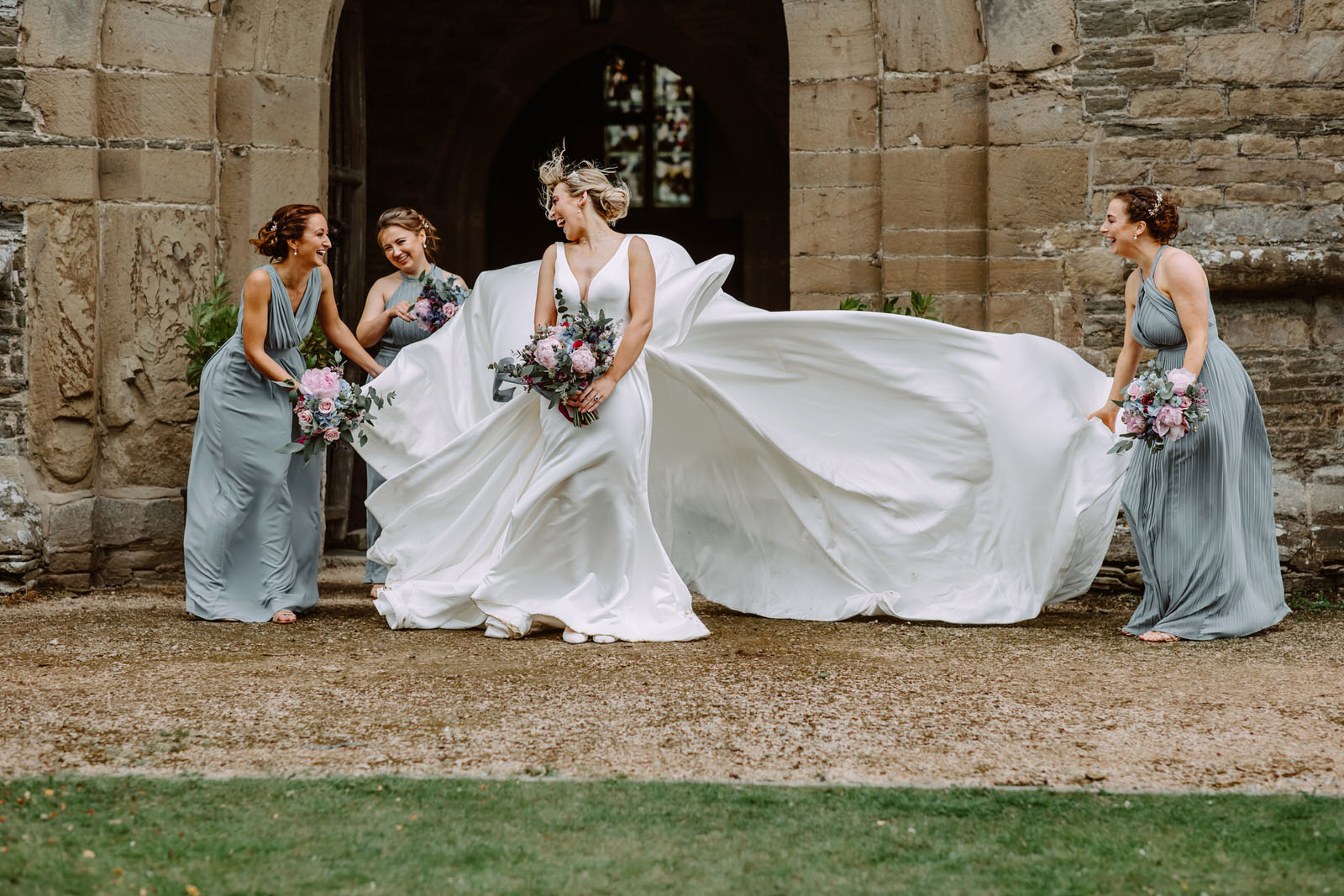 A bride and her bridesmaids holding the wedding dress blown by the wind laughing. 