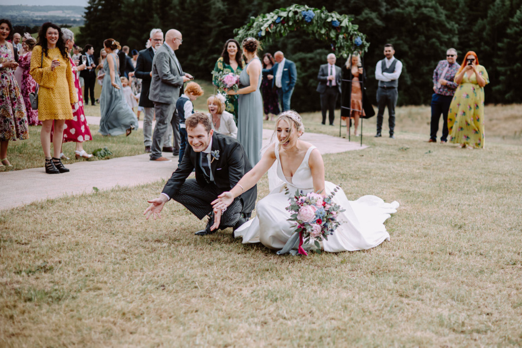 A bride and groom kneeling on the grass in front of their guests.