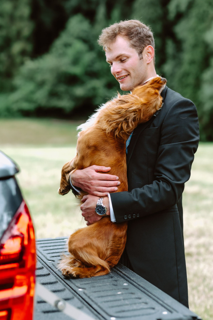 A groom in a suit hugging a dog in the back of a truck.