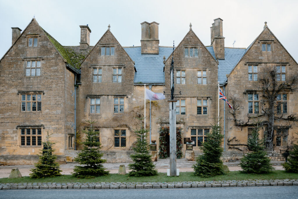 A historic stone building, The  Lygon Arms Hotel, with multiple peaked roofs displaying two flagpoles with flags, is surrounded by small conifer trees along the roadside.