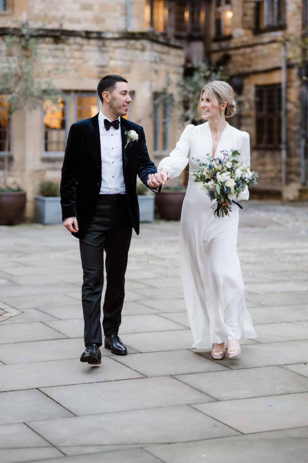 A couple in wedding attire walks hand in hand in a courtyard. The man wears a black tuxedo, and the woman wears a white gown, holding a bouquet. There are stone buildings in the background.