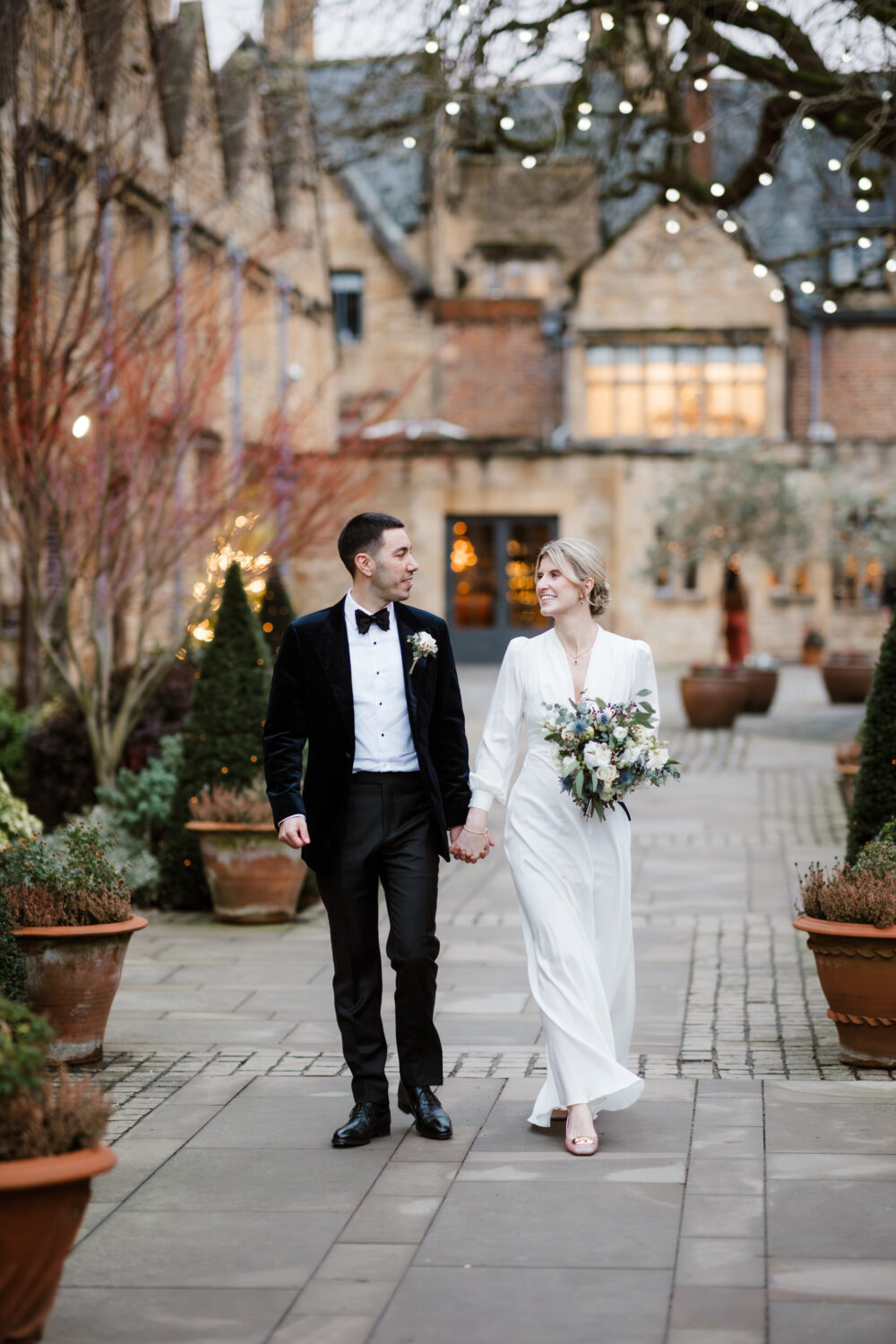 The bride and groom in formal attire walk hand in hand outside a building adorned with string lights. The woman holds a bouquet, and both are smiling, surrounded by potted plants. Cotswolds Wedding Photographer