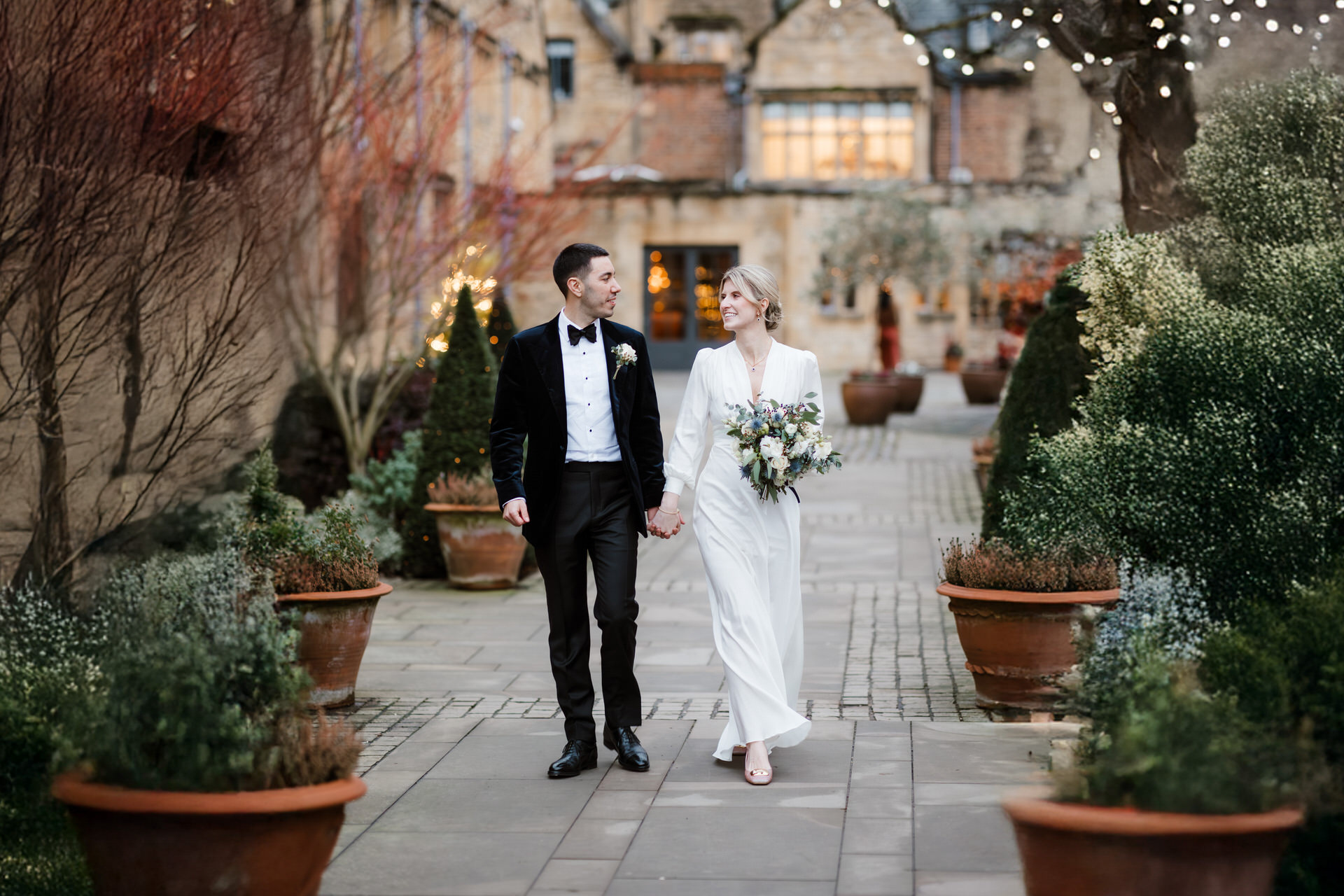 A bride and groom walk hand in hand through a courtyard, surrounded by potted plants and lit trees, wearing a wedding gown and a tuxedo.