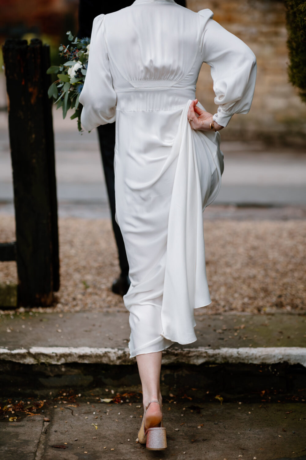 The bride in a white dress holds the hem slightly while walking on a path with a bouquet in one hand.