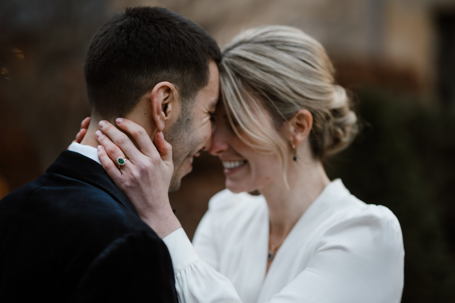 A couple in formal attire embraces closely, smiling with foreheads touching. The woman has her hands on the man's cheeks, while both have eyes closed. The background is softly blurred.