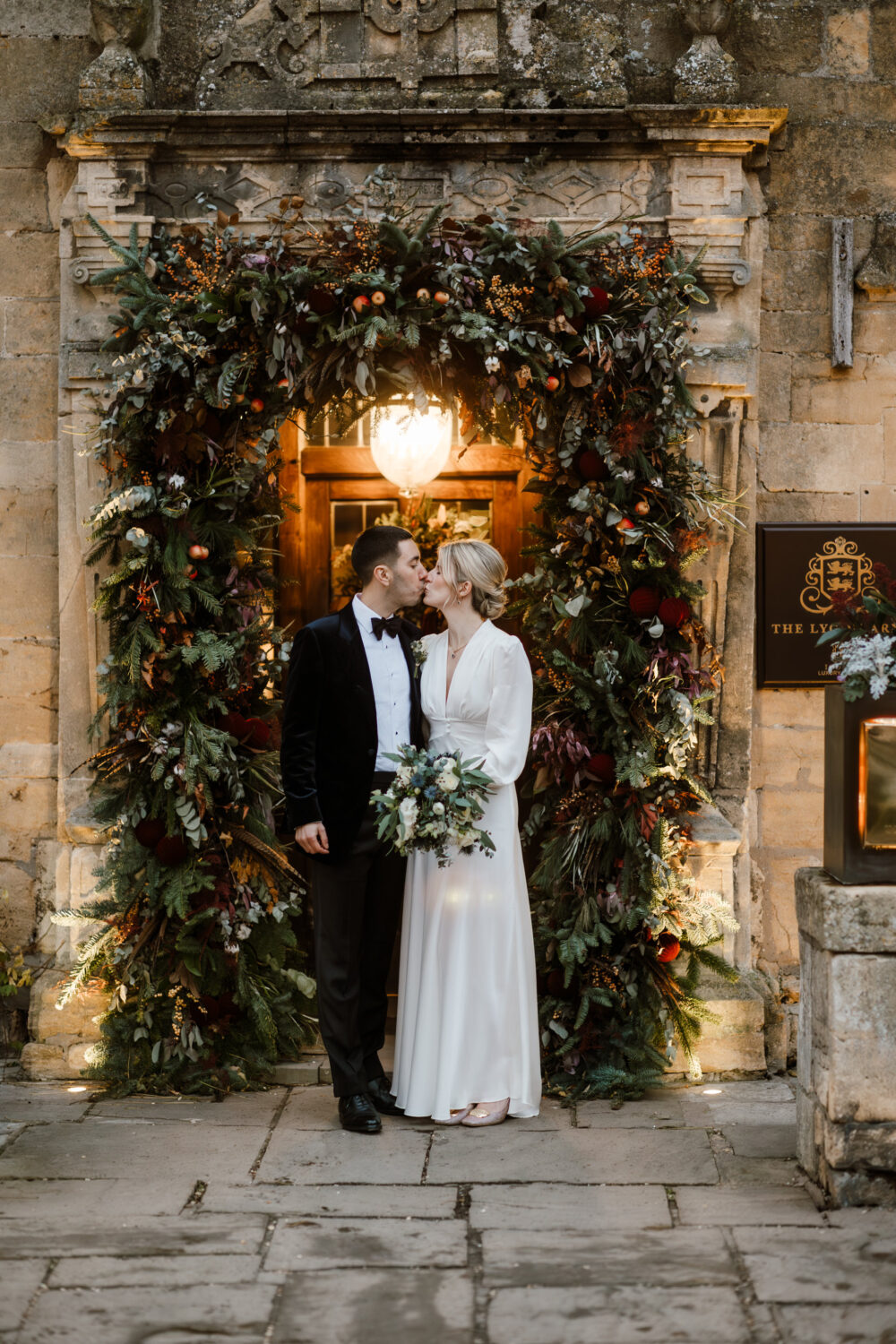 A bride and groom stand in front of a decorative floral archway with greenery. The groom wears a tuxedo, and the bride is in a white dress holding a bouquet.