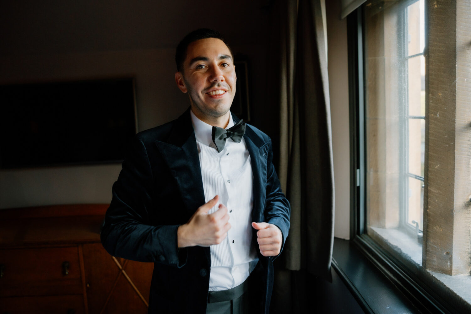 In a formal black velvet tuxedo, the groom stands near a window, adjusting his jacket and smiling.