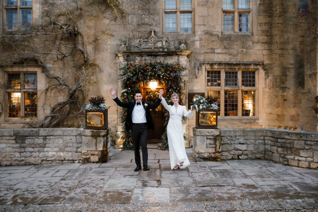 The bride and groom, in formal attire, joyfully wave outside a stone building decorated with foliage and lights. Cotswolds wedding photographer