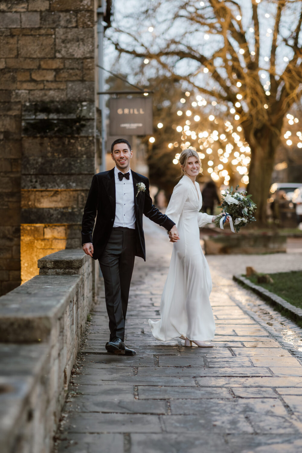 A newlywed couple walks hand in hand on a cobblestone path, with the groom in a tuxedo and the bride in a flowing white dress, set against a backdrop of illuminated trees and a stone building.