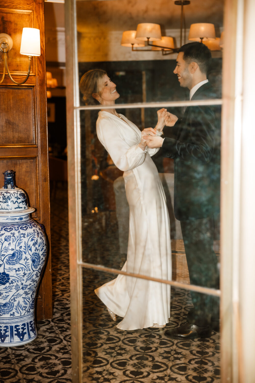 The bride and groom are in an elegant room, viewed through a glass door. The woman is wearing a long white dress, and the man is in a dark suit. A blue and white vase is visible nearby.