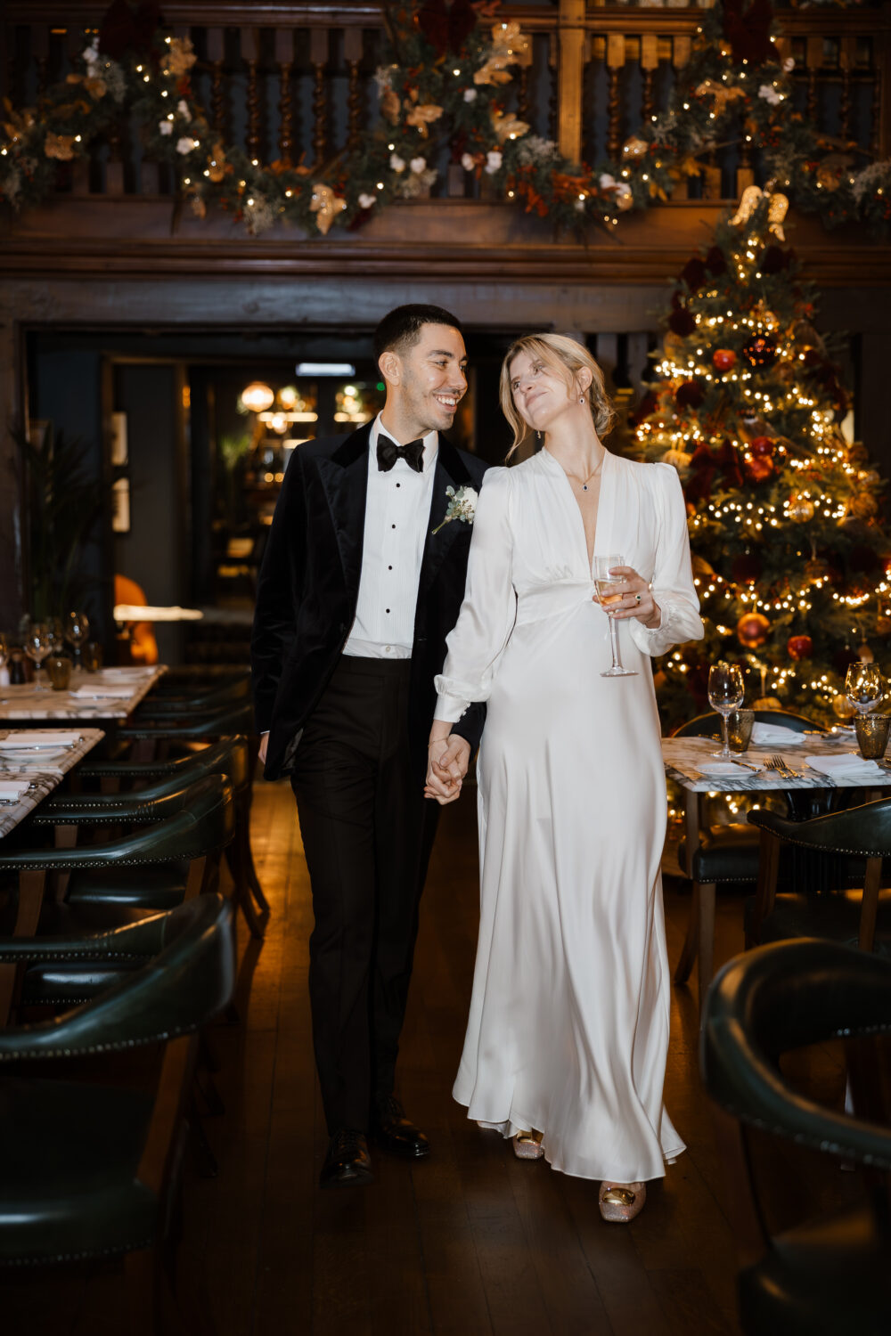 The bride and groom in formal attire, the woman in a white dress holding a glass, and the man in a tuxedo, walk hand in hand in a festive setting with a lit Christmas tree.