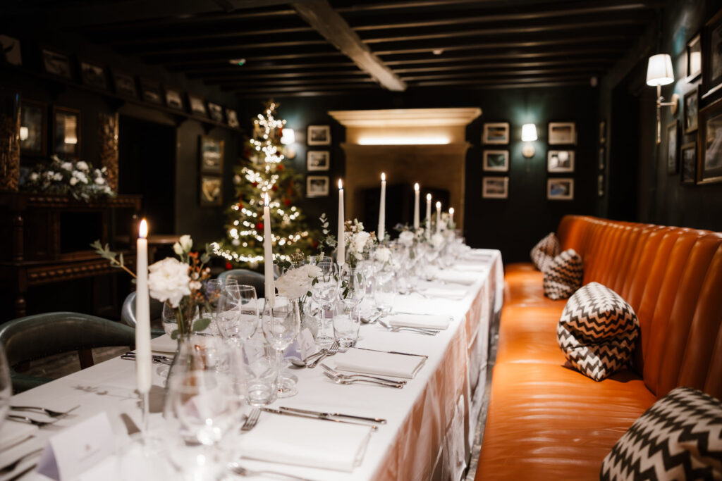 A long dining table is elegantly set with white linens, candles, and glassware. A Christmas tree is in the background and a cushioned bench with patterned pillows lines one side. Wedding breakfast