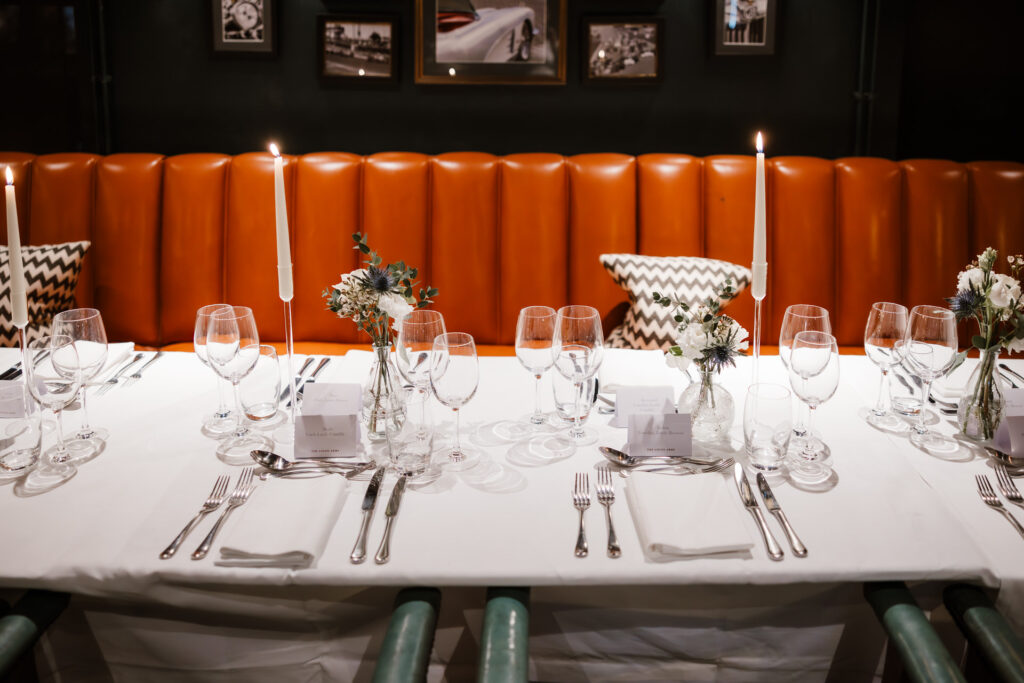 A table set for a formal dinner with white tablecloth, wine glasses, silver cutlery, and floral arrangements. Tall candles are lit on either side. Orange cushioned seating in the background.