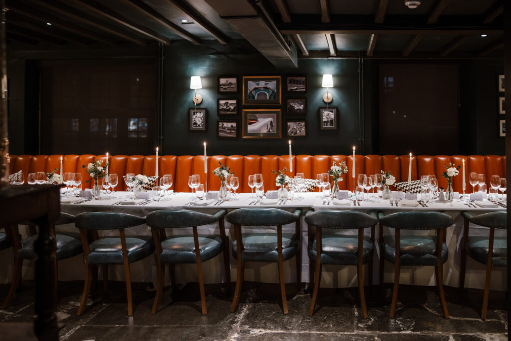 An elegant dining room with a long table featuring candles and flowers. Orange banquette seating against dark walls adorned with framed pictures ready for wedding breakfast