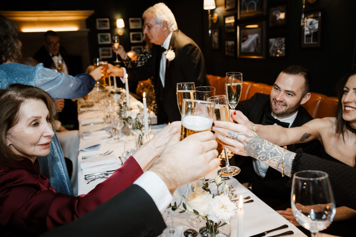 Group of people seated at a dinner table raising glasses for a toast, with a floral centerpiece and framed pictures on a dark green wall in the background.