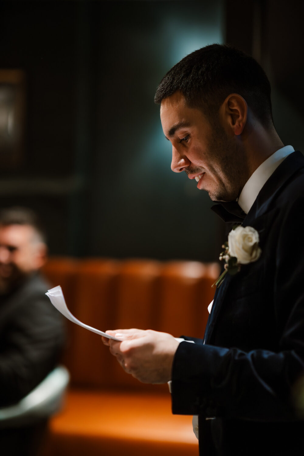 The groom is in a suit, holding a paper, reading, or giving a speech indoors. Another person is blurred in the background.