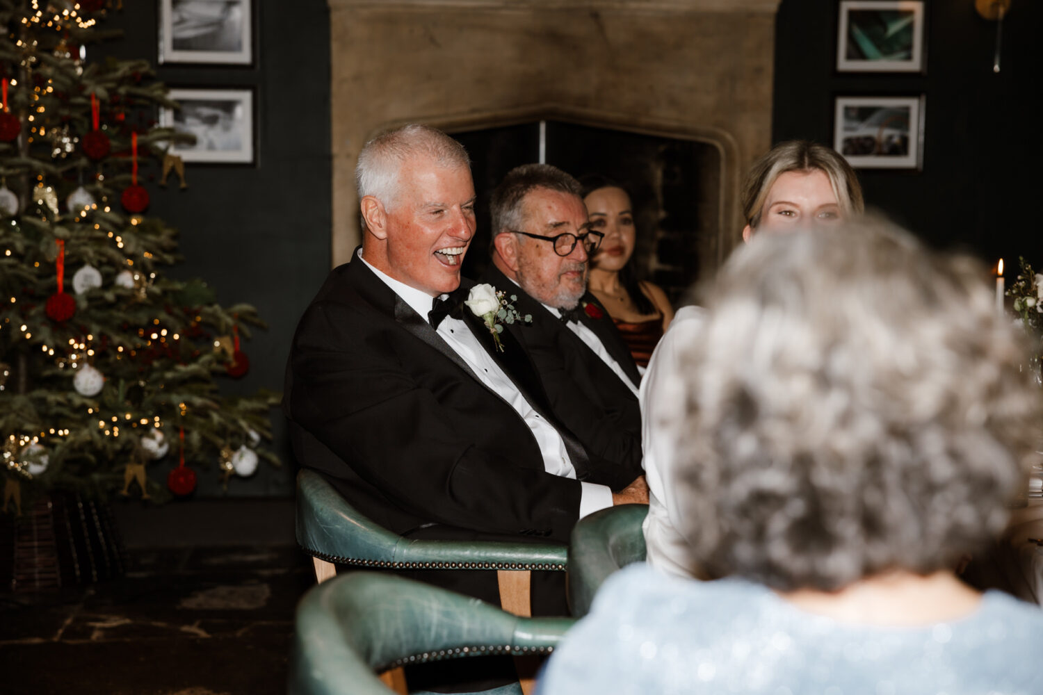 A group of people in formal attire are sitting around a table. A decorated Christmas tree is visible in the background.