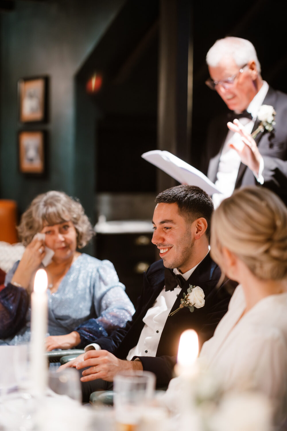 A man in a tuxedo gives a speech at a formal dinner. A man and a woman sit and listen, with candles and blurred background pictures. Another woman wipes her eye with a tissue.