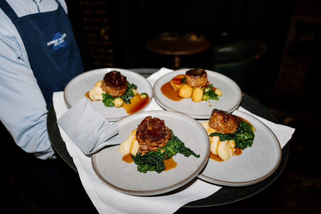 A server carries four plates of beef medallions on mashed potatoes with greens and sauce.