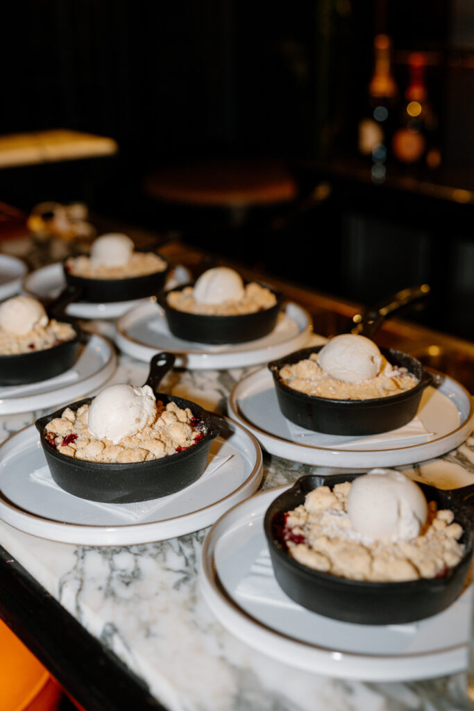 Small cast iron skillets filled with crisp dessert topped with a scoop of vanilla ice cream, arranged on white plates on a marble countertop.