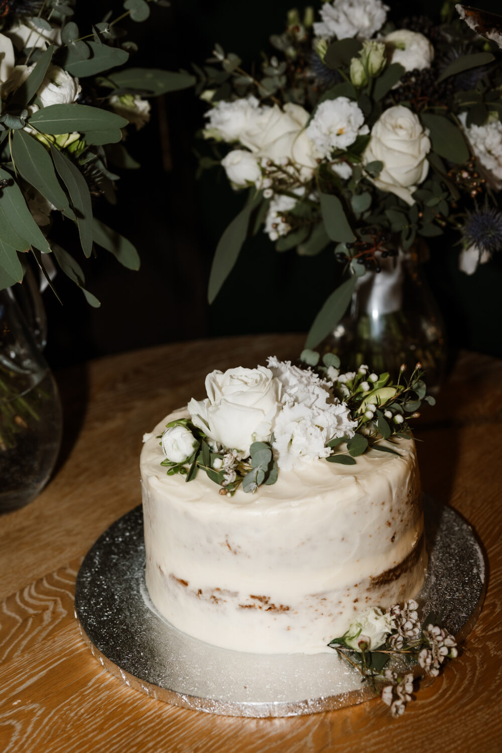 A two-tiered wedding cake with white frosting decorated with white roses and greenery sits on a silver platter. Two floral arrangements are visible in the background.