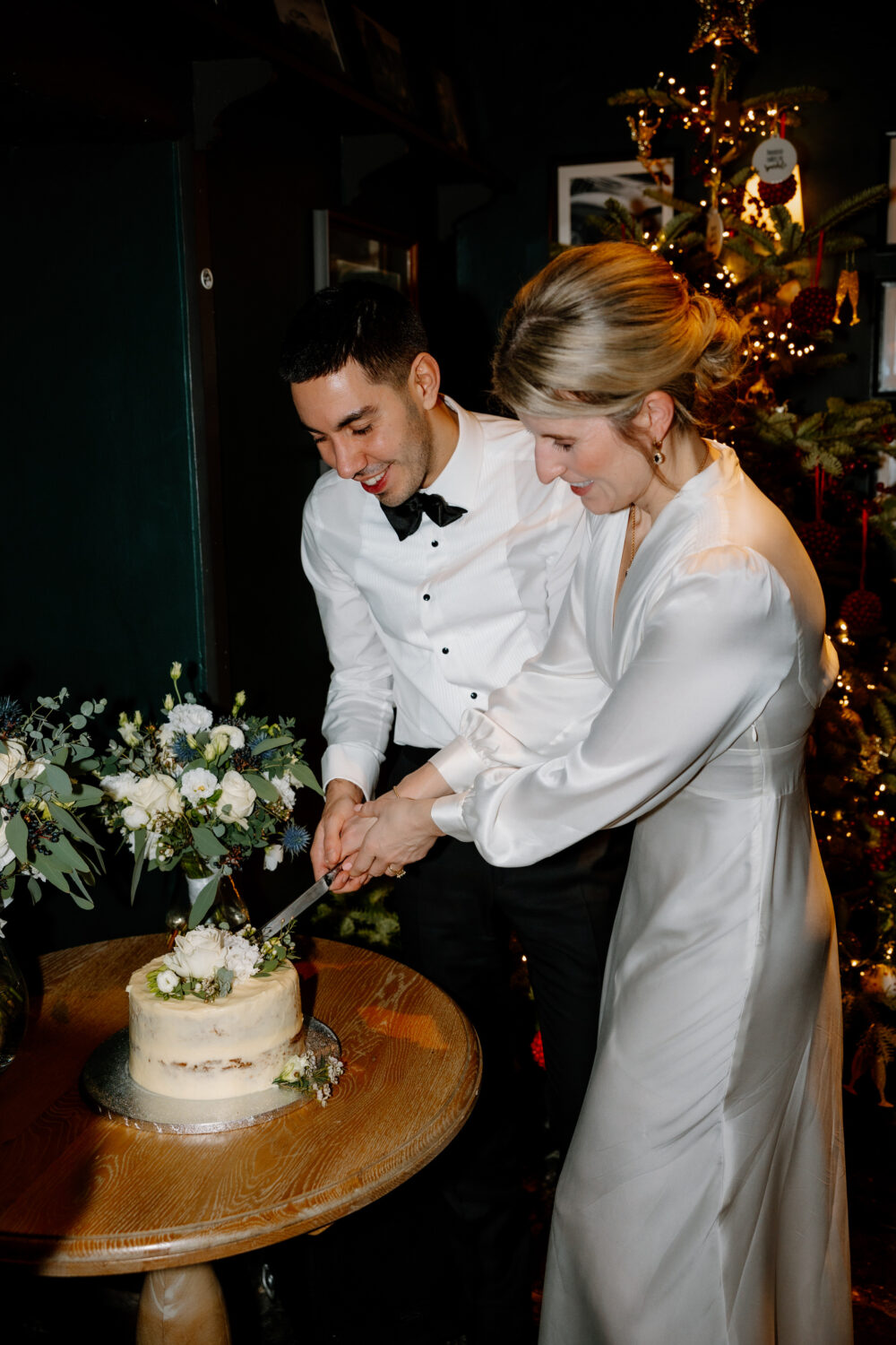 A man and woman in formal attire cut a small wedding cake together. A decorated Christmas tree is in the background.