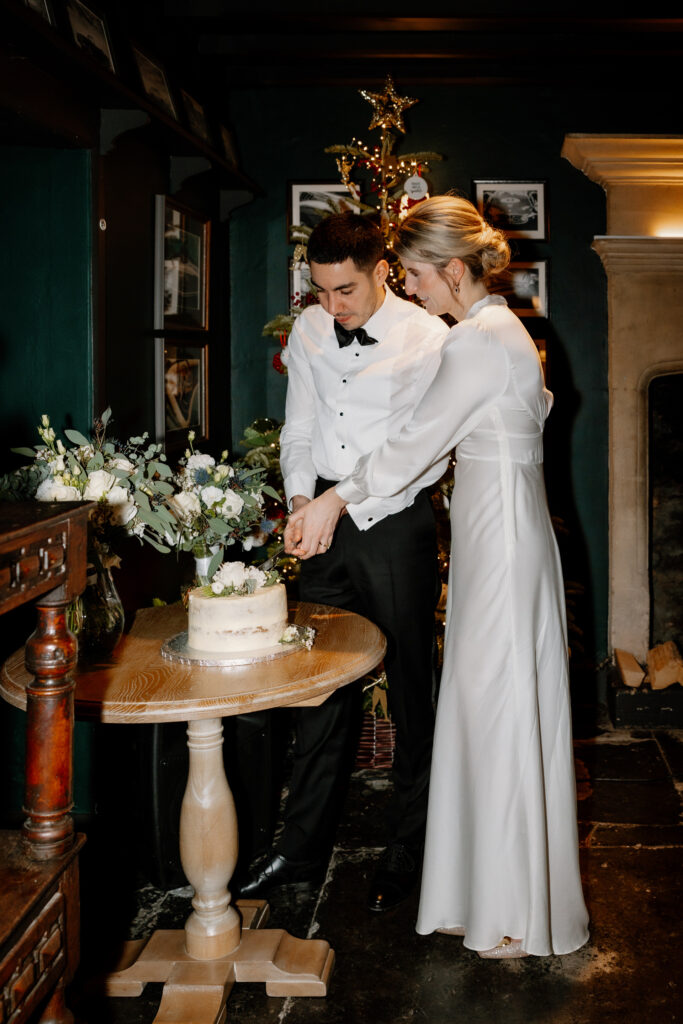 A couple in formal attire cuts a cake on a small round table. A decorative plant and framed photos are in the background.