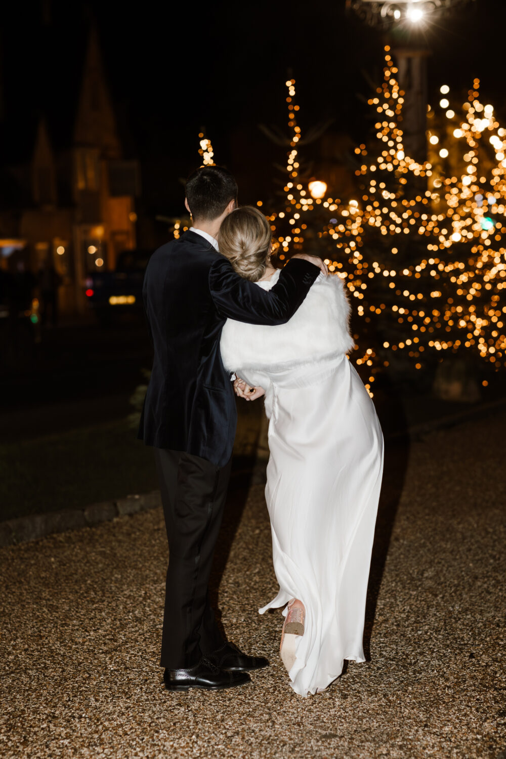 A couple in formal attire, facing away, stand together outside at night near lit Christmas trees.