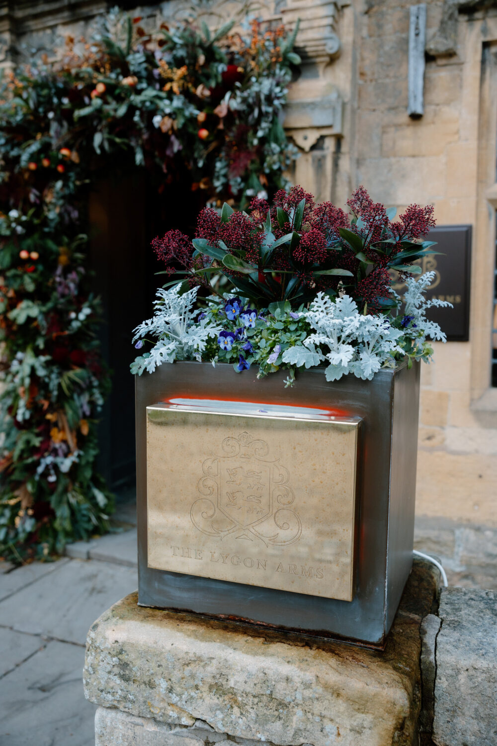 A metal planter with a crest, filled with green and purple foliage and white flowers, set in front of a stone building with an arched floral entrance.