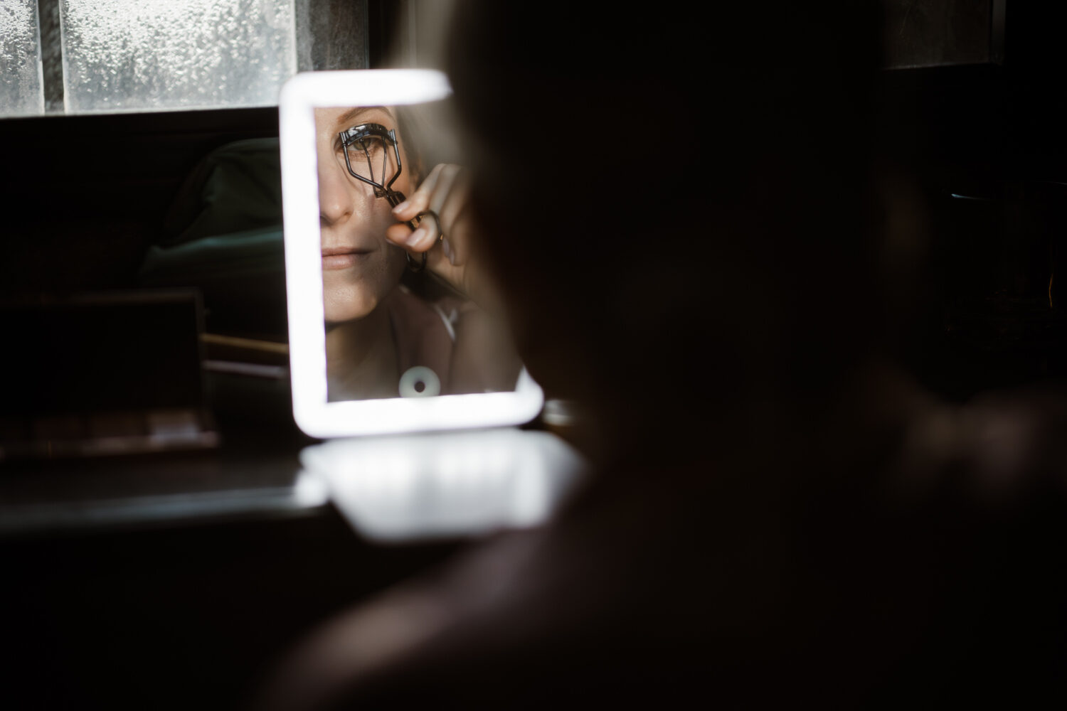 The bride uses an eyelash curler in front of an illuminated mirror with a dimly lit background.