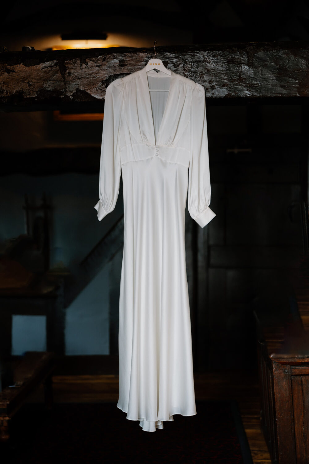 A long-sleeved, white satin wedding dress hangs from a wooden beam in a dimly lit room.