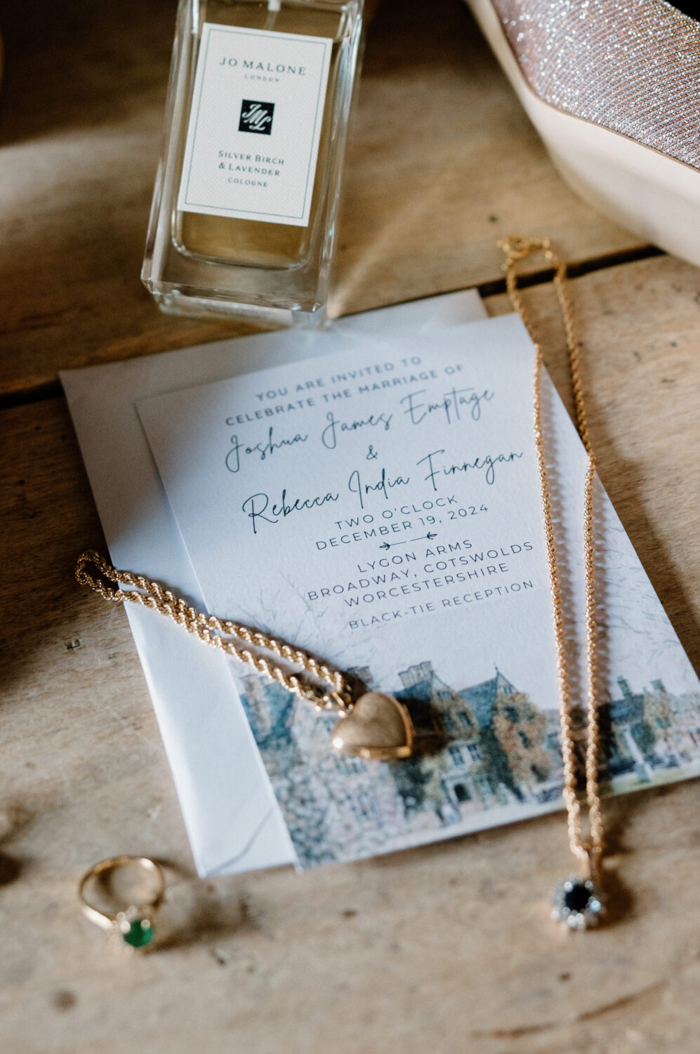 Invitation card for a wedding ceremony, a bottle of Jo Malone perfume, gold necklaces, and a ring with a green gemstone arranged on a wooden surface.